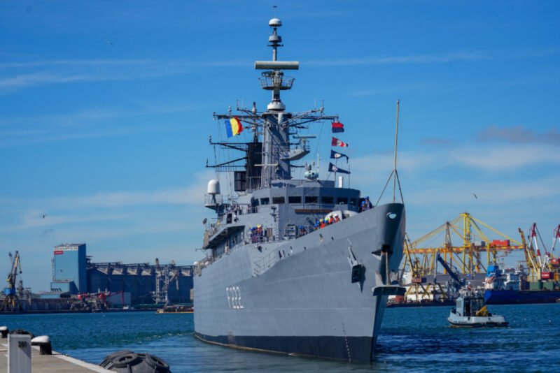 Gray naval destroyer docked at a harbor with cranes and a bright blue sky behind it, flags on the mast visible at the bow.