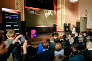 Audience watching a formal stage presentation in an ornate theater, with a speaker at a podium and large display screens on stage.