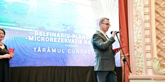 Man in a suit speaks into a microphone on a stage, with a large blue display behind and a woman holding a red folder to the left.