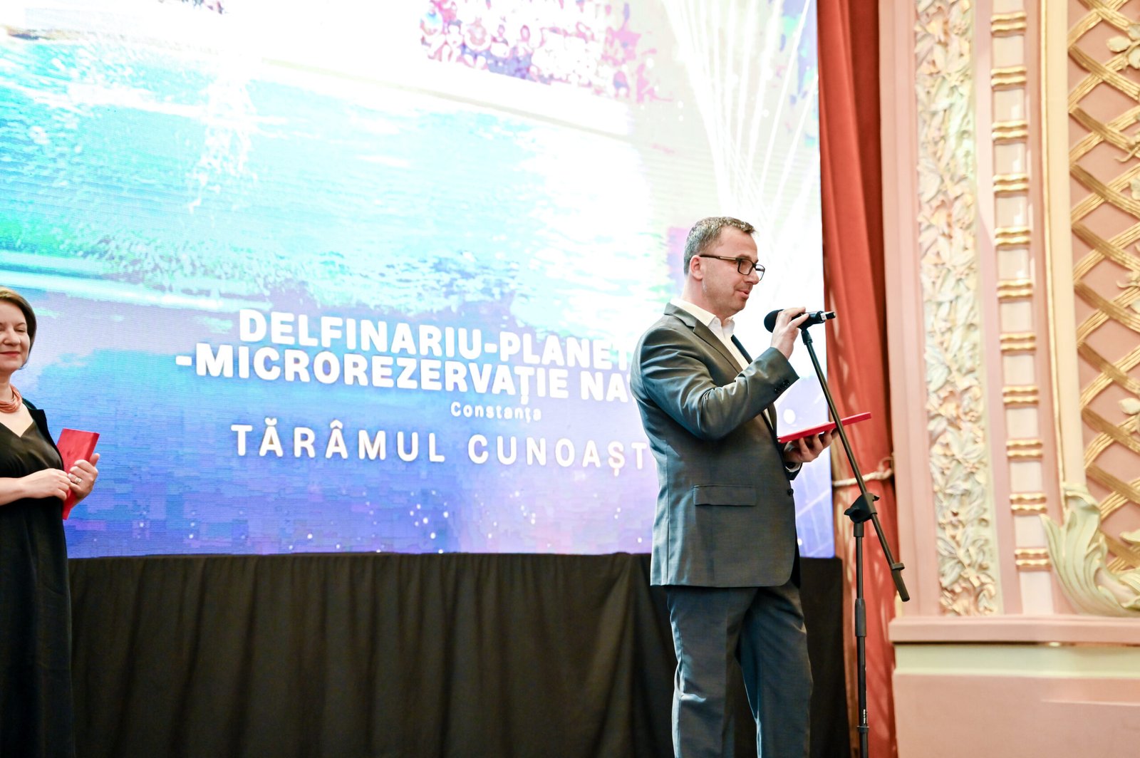 Man in a suit speaks into a microphone on a stage, with a large blue display behind and a woman holding a red folder to the left.