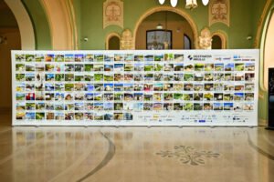 Large white display board filled with dozens of travel photos and logos, set in an ornate green-walled hall.