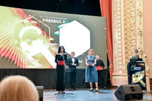 Group of presenters on a stage holding red folders, with a large LED screen and ornate theater decor on the right.