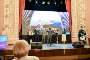 Presenter speaks into a microphone on a grand theater stage while several award recipients stand with plaques behind him, with a large LED screen backdrop behind them and ornate side walls visible.