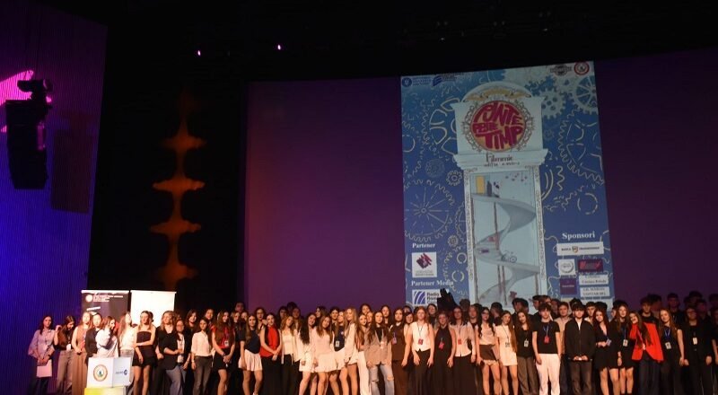 Large group of young people on a theater stage posing for a group photo during an event, with a tall blue graphic poster projected on a screen behind them and red balloons in the foreground.