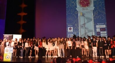 Large group of young people on a theater stage posing for a group photo during an event, with a tall blue graphic poster projected on a screen behind them and red balloons in the foreground.