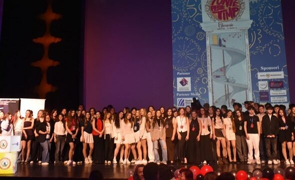 Large group of young people on a theater stage posing for a group photo during an event, with a tall blue graphic poster projected on a screen behind them and red balloons in the foreground.