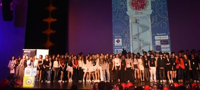 Large group of young people on a theater stage posing for a group photo during an event, with a tall blue graphic poster projected on a screen behind them and red balloons in the foreground.