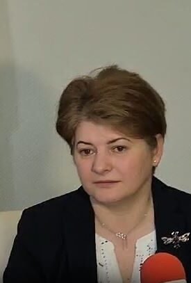 Woman in a dark blazer speaks into a red microphone during an interview, with flags in the background.