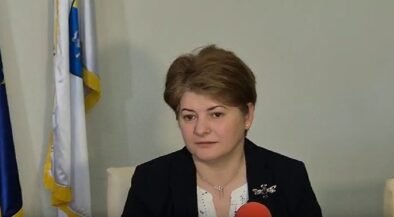 Woman in a dark blazer speaks into a red microphone during an interview, with flags in the background.