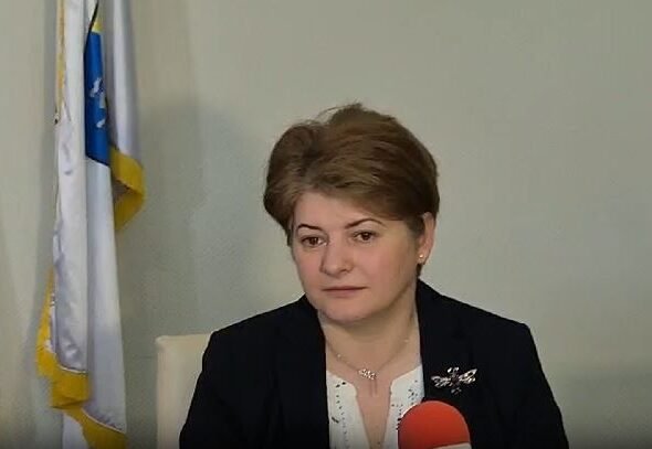 Woman in a dark blazer speaks into a red microphone during an interview, with flags in the background.