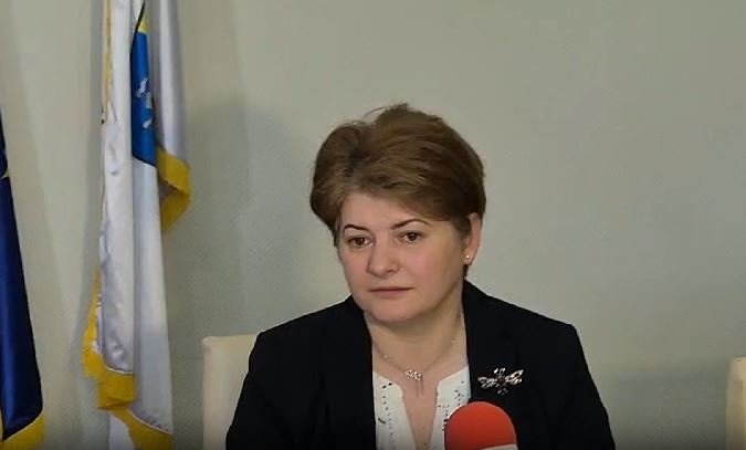 Woman in a dark blazer speaks into a red microphone during an interview, with flags in the background.