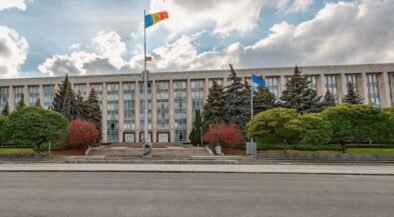 Front of a large government building with steps, flagpoles, and manicured trees in front of it in an urban plaza.