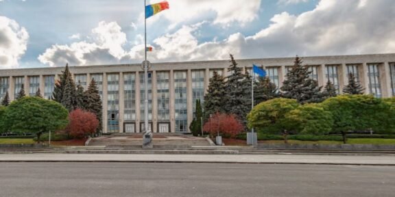 Front of a large government building with steps, flagpoles, and manicured trees in front of it in an urban plaza.