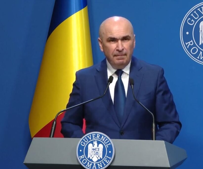 A bald man in a dark blue suit and tie speaking at a podium with two microphones, a Ukrainian flag behind him, and a blue backdrop with a circular emblem.