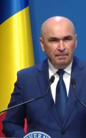 A bald man in a dark blue suit and tie speaking at a podium with two microphones, a Ukrainian flag behind him, and a blue backdrop with a circular emblem.