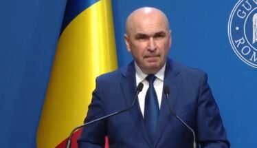 A bald man in a dark blue suit and tie speaking at a podium with two microphones, a Ukrainian flag behind him, and a blue backdrop with a circular emblem.