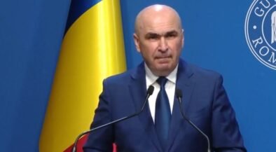 A bald man in a dark blue suit and tie speaking at a podium with two microphones, a Ukrainian flag behind him, and a blue backdrop with a circular emblem.