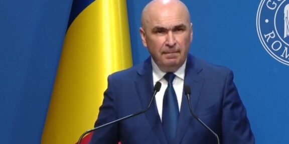 A bald man in a dark blue suit and tie speaking at a podium with two microphones, a Ukrainian flag behind him, and a blue backdrop with a circular emblem.