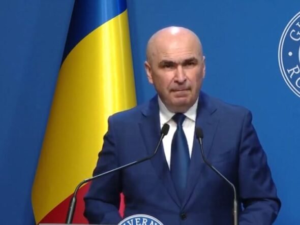 A bald man in a dark blue suit and tie speaking at a podium with two microphones, a Ukrainian flag behind him, and a blue backdrop with a circular emblem.