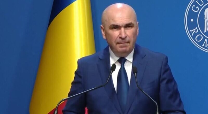 A bald man in a dark blue suit and tie speaking at a podium with two microphones, a Ukrainian flag behind him, and a blue backdrop with a circular emblem.