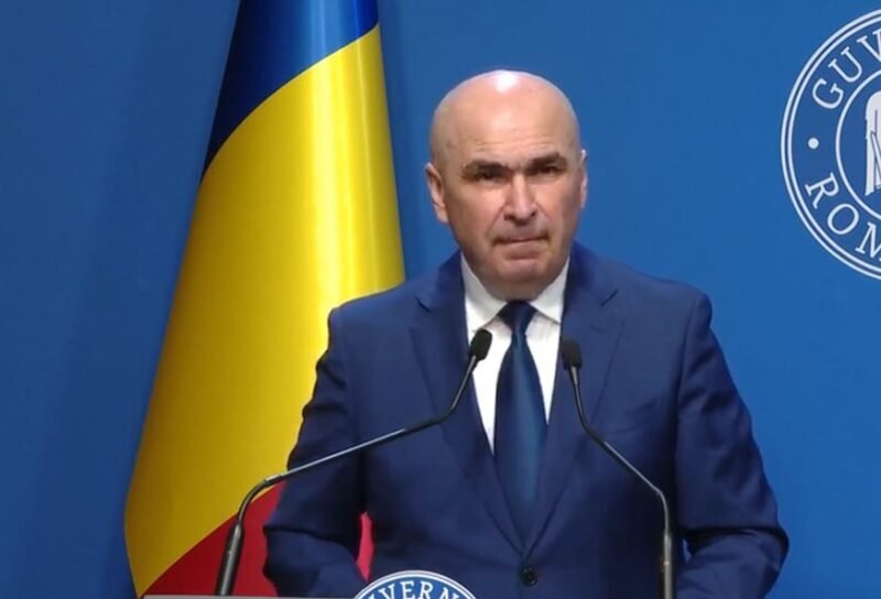 A bald man in a dark blue suit and tie speaking at a podium with two microphones, a Ukrainian flag behind him, and a blue backdrop with a circular emblem.