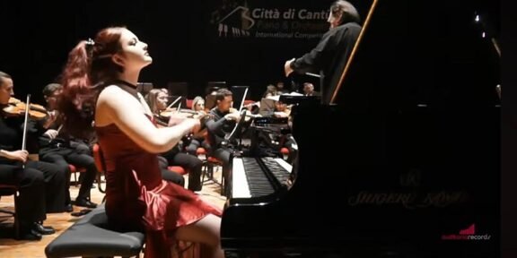 Female pianist in a red dress playing a grand piano on stage, an orchestra and conductor visible in the background.