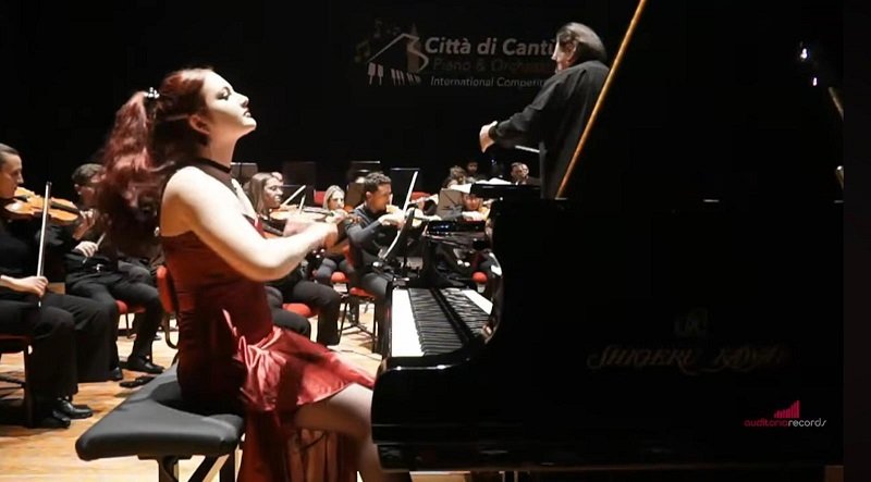 Female pianist in a red dress playing a grand piano on stage, an orchestra and conductor visible in the background.