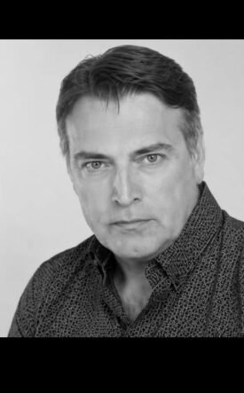 Black-and-white head-and-shoulders portrait of a man with dark hair wearing a patterned shirt, looking at the camera in a neutral expression in a studio setting.