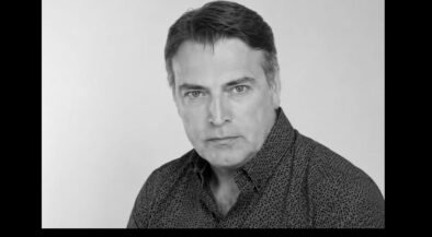 Black-and-white head-and-shoulders portrait of a man with dark hair wearing a patterned shirt, looking at the camera in a neutral expression in a studio setting.