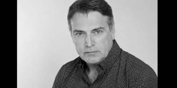 Black-and-white head-and-shoulders portrait of a man with dark hair wearing a patterned shirt, looking at the camera in a neutral expression in a studio setting.