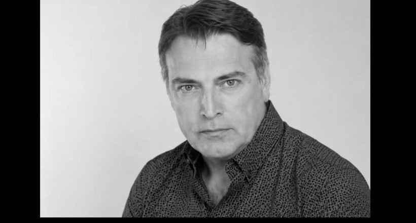 Black-and-white head-and-shoulders portrait of a man with dark hair wearing a patterned shirt, looking at the camera in a neutral expression in a studio setting.
