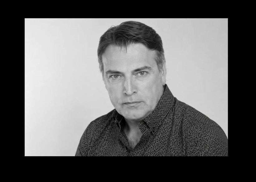 Black-and-white head-and-shoulders portrait of a man with dark hair wearing a patterned shirt, looking at the camera in a neutral expression in a studio setting.