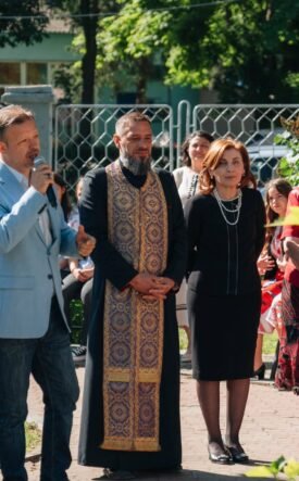 Man in light blue blazer speaks into a microphone at an outdoor ceremony, surrounded by adults in formal attire and traditional clothing.