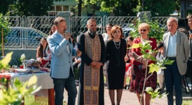 Man in light blue blazer speaks into a microphone at an outdoor ceremony, surrounded by adults in formal attire and traditional clothing.