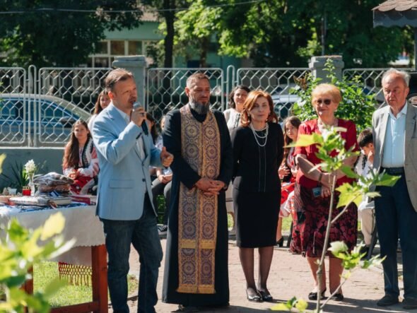 Man in light blue blazer speaks into a microphone at an outdoor ceremony, surrounded by adults in formal attire and traditional clothing.