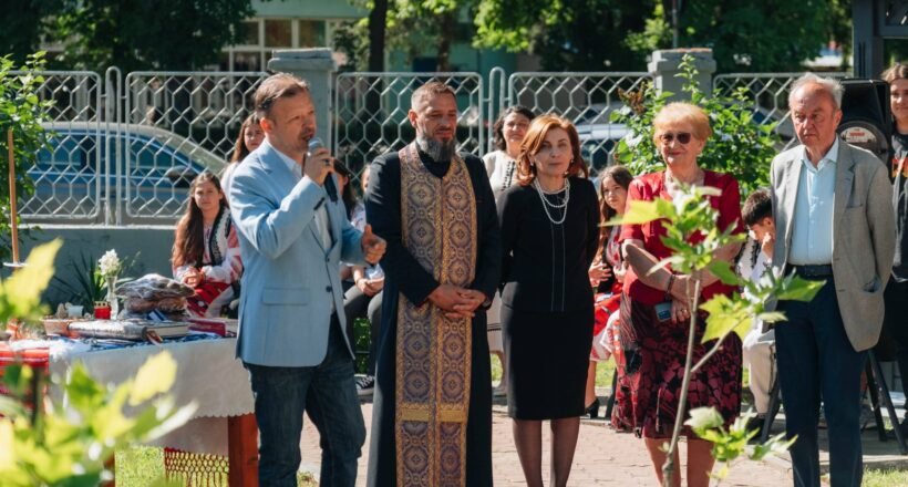 Man in light blue blazer speaks into a microphone at an outdoor ceremony, surrounded by adults in formal attire and traditional clothing.