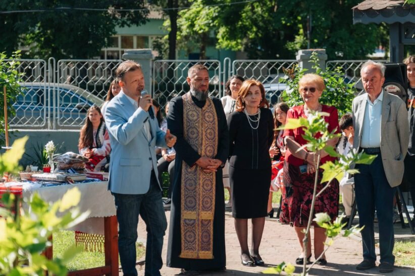 Man in light blue blazer speaks into a microphone at an outdoor ceremony, surrounded by adults in formal attire and traditional clothing.