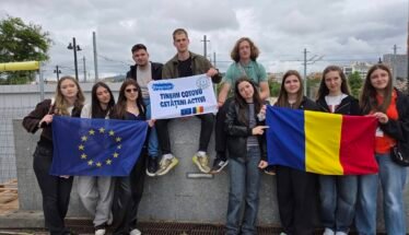 Group of diverse young students on a rooftop, holding the EU flag and the Romanian flag with an Erasmus banner saying active citizens.