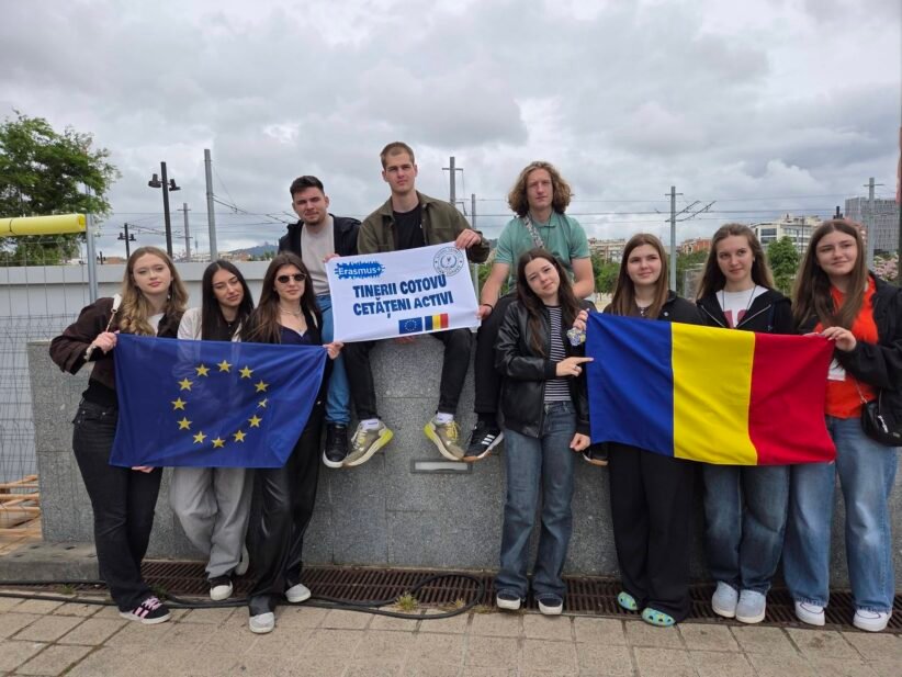 Group of diverse young students on a rooftop, holding the EU flag and the Romanian flag with an Erasmus banner saying active citizens.