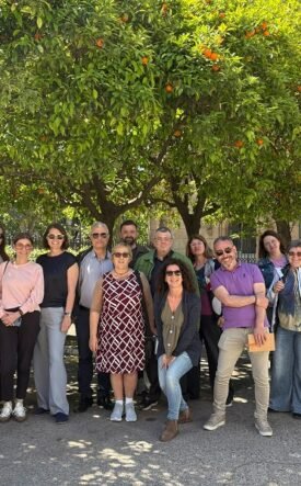 Group of smiling adults posing together outdoors under orange trees in a sunny courtyard with a historic stone building in the background