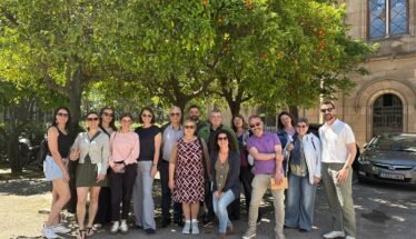 Group of smiling adults posing together outdoors under orange trees in a sunny courtyard with a historic stone building in the background