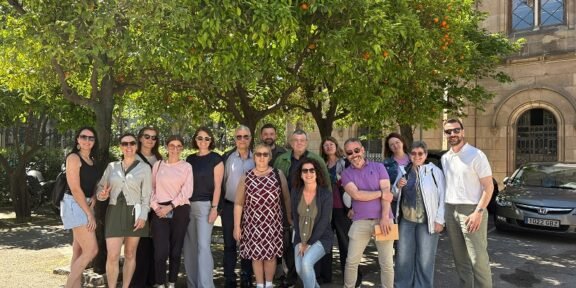 Group of smiling adults posing together outdoors under orange trees in a sunny courtyard with a historic stone building in the background
