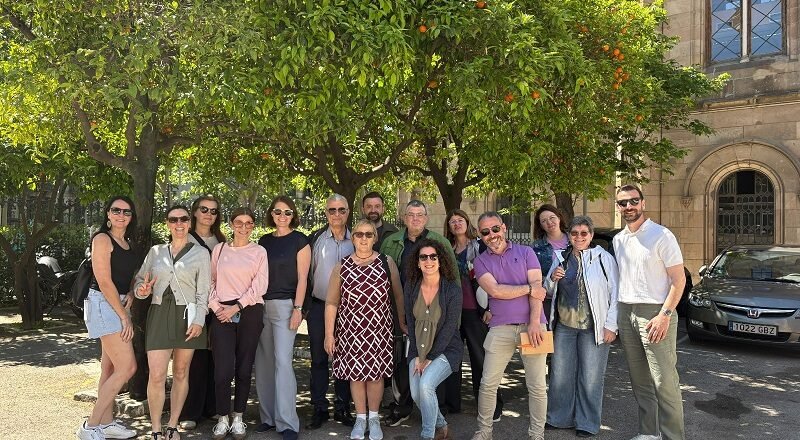Group of smiling adults posing together outdoors under orange trees in a sunny courtyard with a historic stone building in the background