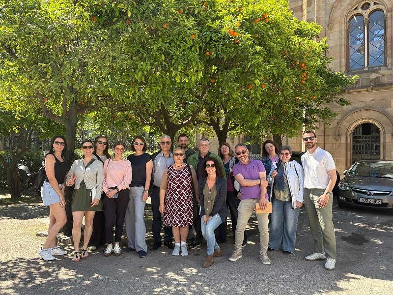 Group of smiling adults posing together outdoors under orange trees in a sunny courtyard with a historic stone building in the background
