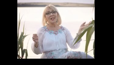 Older woman with blonde hair and glasses sits by a lake, wearing a white embroidered blouse and turquoise accessories, speaking or singing to the camera.