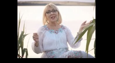 Older woman with blonde hair and glasses sits by a lake, wearing a white embroidered blouse and turquoise accessories, speaking or singing to the camera.