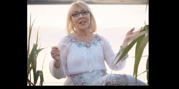 Older woman with blonde hair and glasses sits by a lake, wearing a white embroidered blouse and turquoise accessories, speaking or singing to the camera.