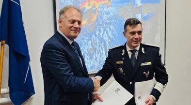 Two men shake hands in a formal room; one in a suit and the other in a military uniform with medals, holding documents behind them with a projector screen the background showing helicopters and a flag on the left side.