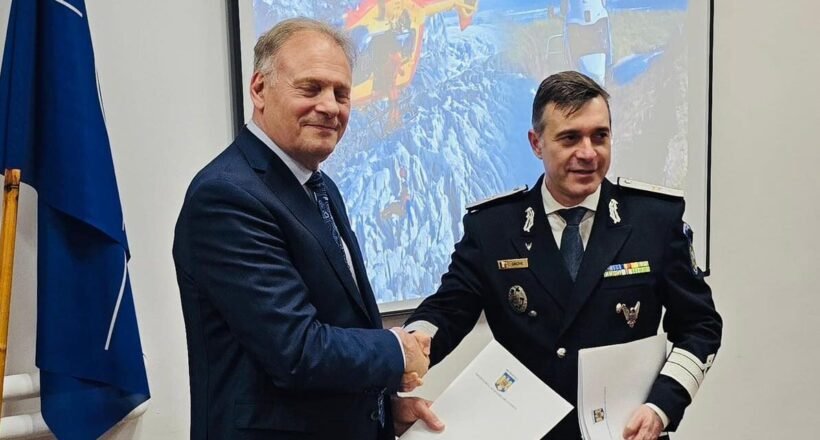 Two men shake hands in a formal room; one in a suit and the other in a military uniform with medals, holding documents behind them with a projector screen the background showing helicopters and a flag on the left side.