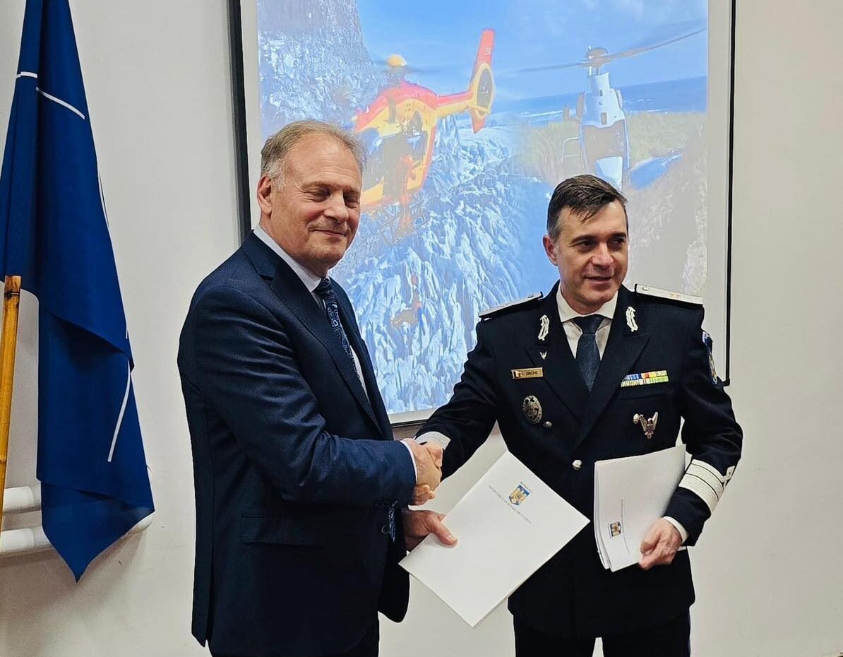 Two men shake hands in a formal room; one in a suit and the other in a military uniform with medals, holding documents behind them with a projector screen the background showing helicopters and a flag on the left side.